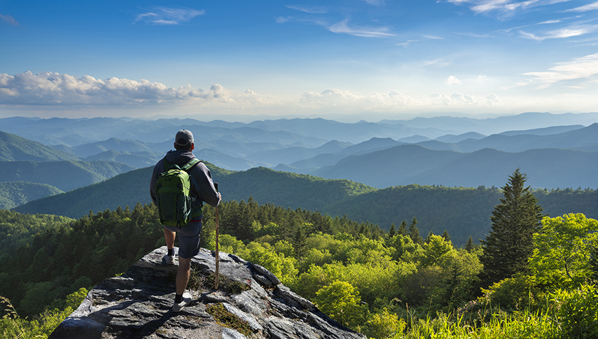 A hiker with a green backpack stands on a rocky cliff, overlooking a mountainous valley