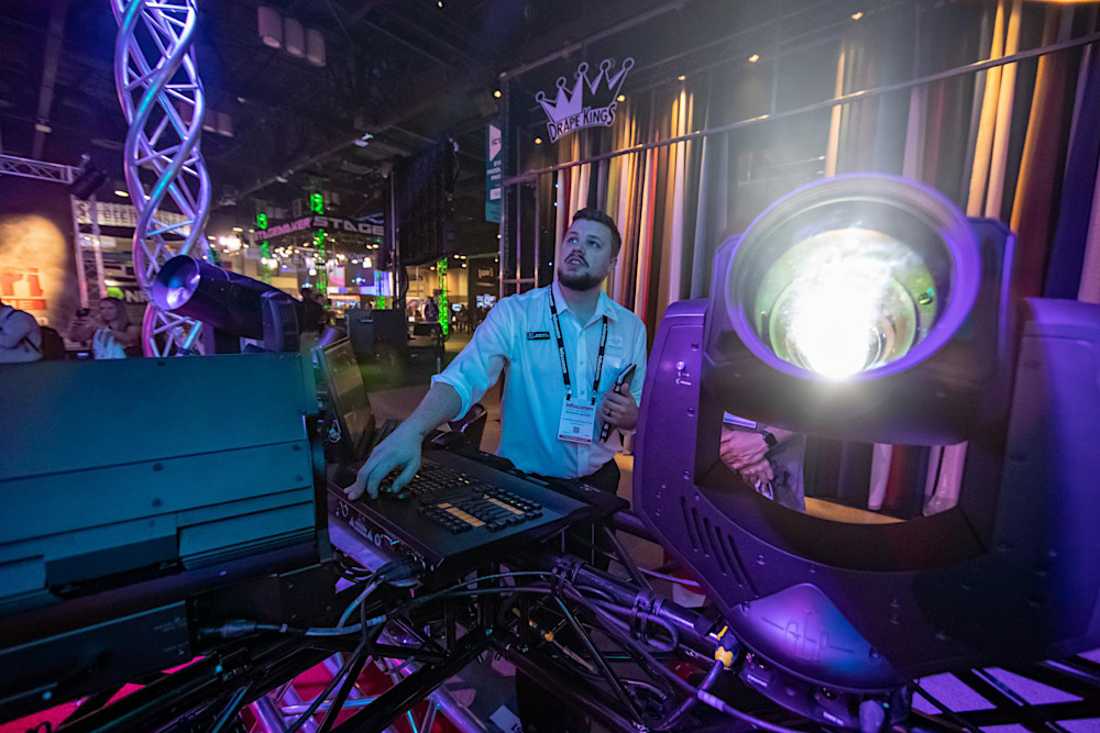 A man operates stage lighting equipment at an event, surrounded by colorful lights and a backdrop of curtains.