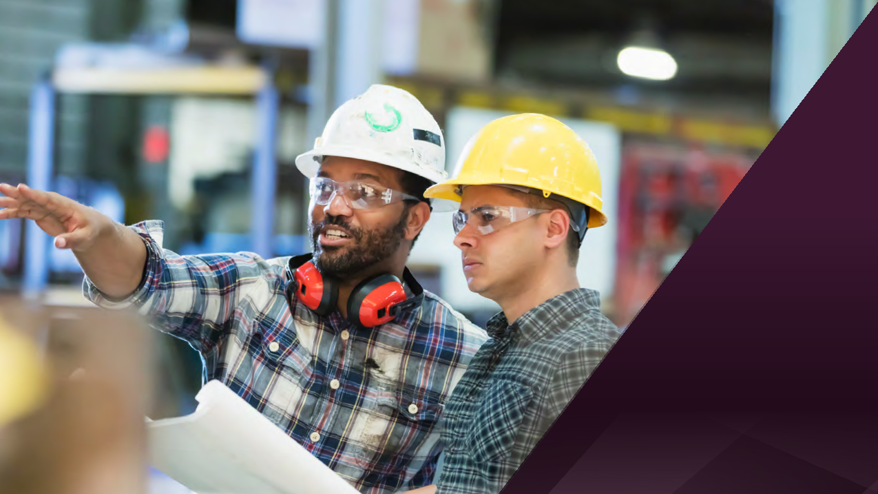 Two workers in plaid shirts and hard hats, discussing plans in an industrial setting. One points forward, holding a blueprint.