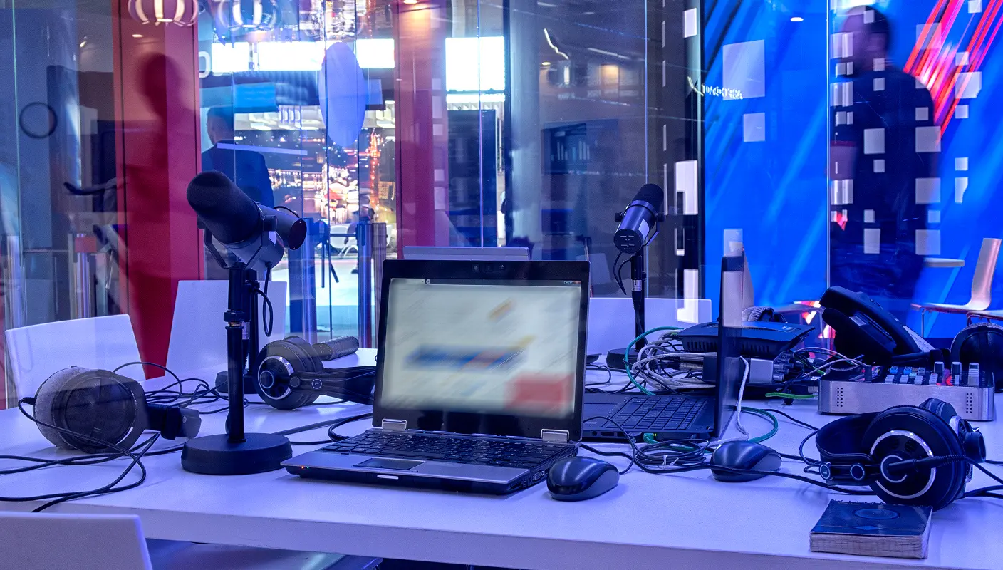 A radio studio setup with microphones, headphones, a mixer, and a laptop on a desk in a modern, colorful environment.