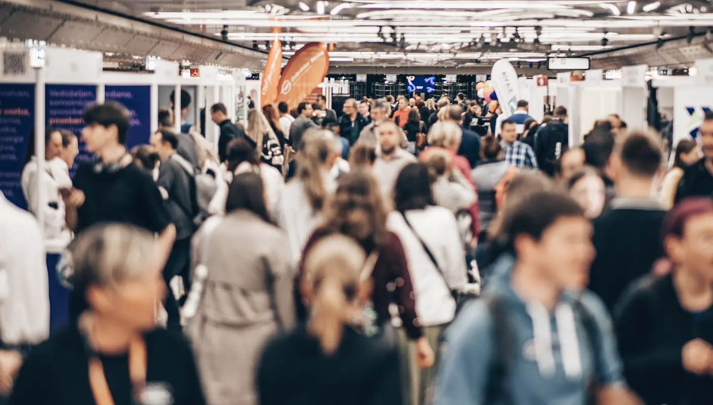 Crowded exhibition hall with people walking and visiting booths, some holding informational materials. Bright lighting and banners are visible.