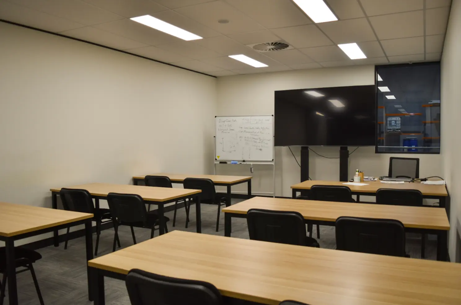 Empty classroom with wooden desks and black chairs, a whiteboard, and a large TV screen on the wall.