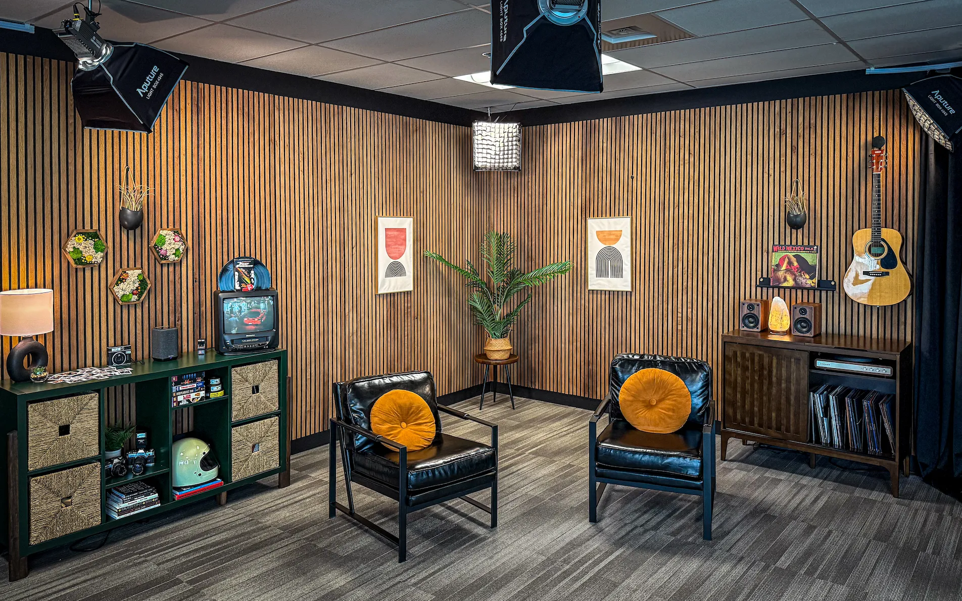 Cozy room with wood paneling, two chairs with orange cushions, wall art, a guitar, shelf with decor, and overhead studio lights.