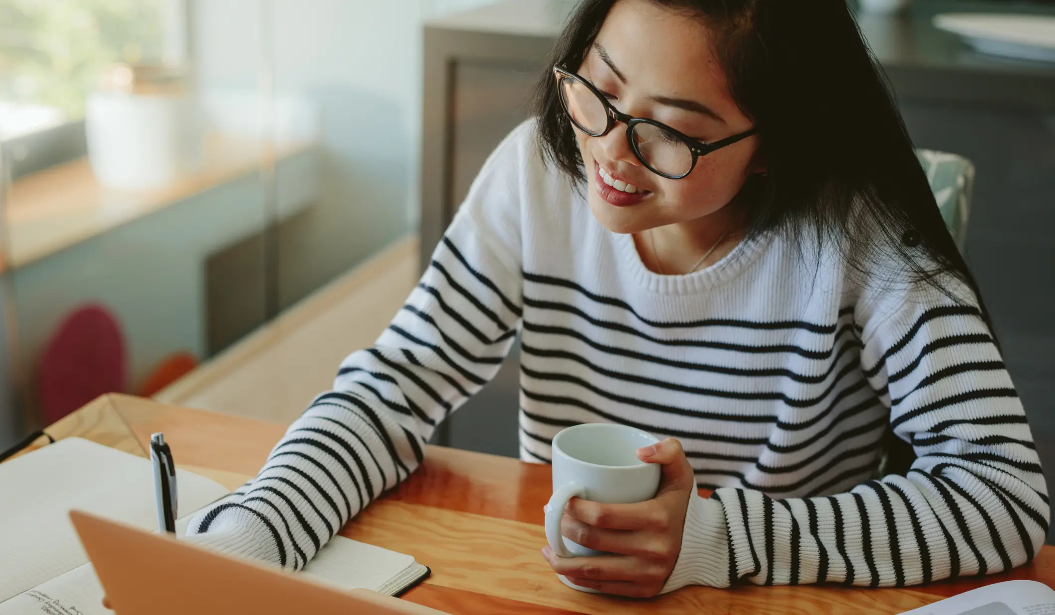 A woman in a striped sweater, wearing glasses, writes in a notebook while holding a mug, seated by a laptop on a wooden desk.