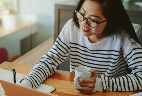 A woman in a striped sweater, wearing glasses, writes in a notebook while holding a mug, seated by a laptop on a wooden desk.