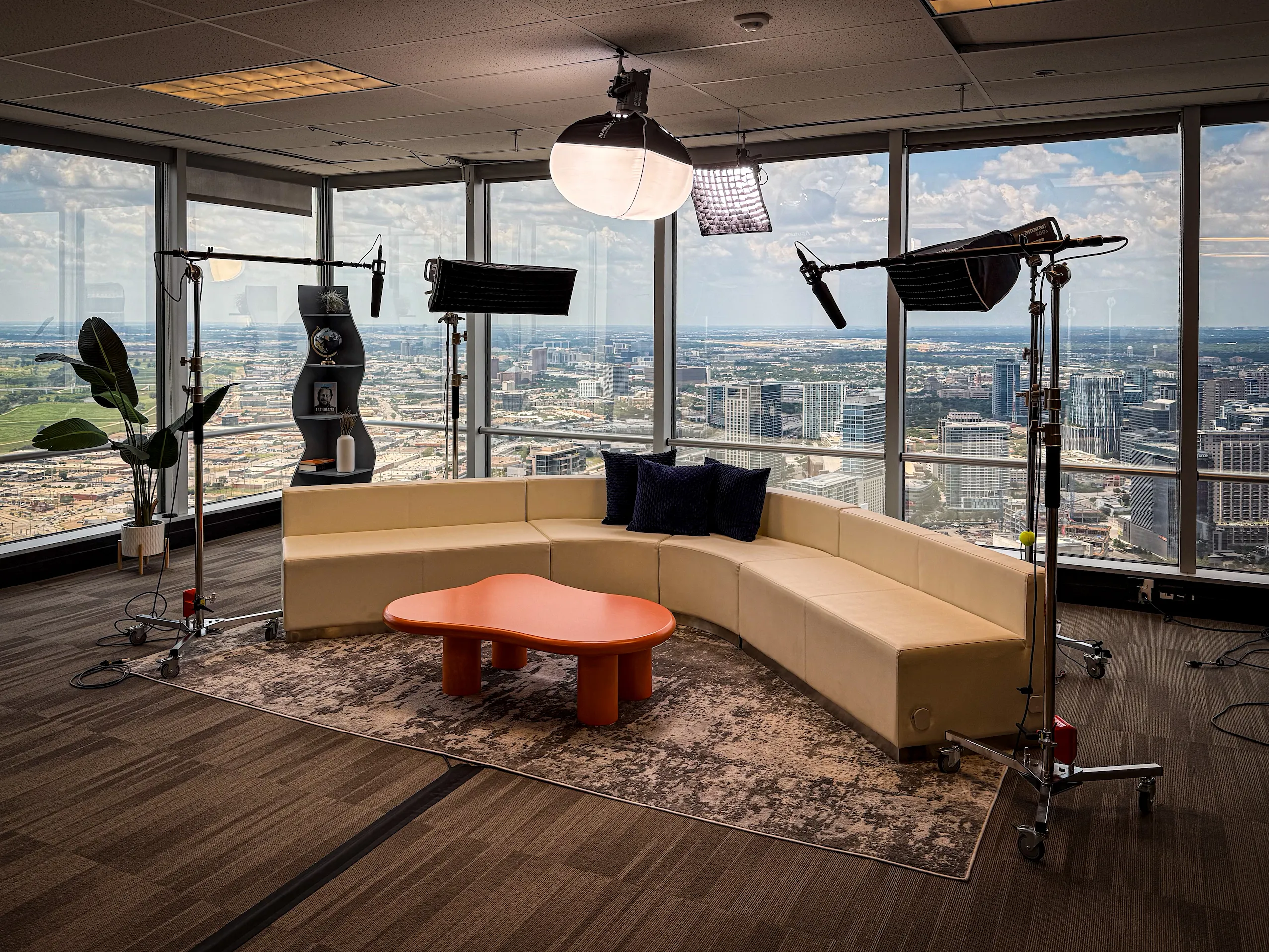 Modern office corner setup with beige sectional sofa, orange table, studio lights, and city view through large windows.
