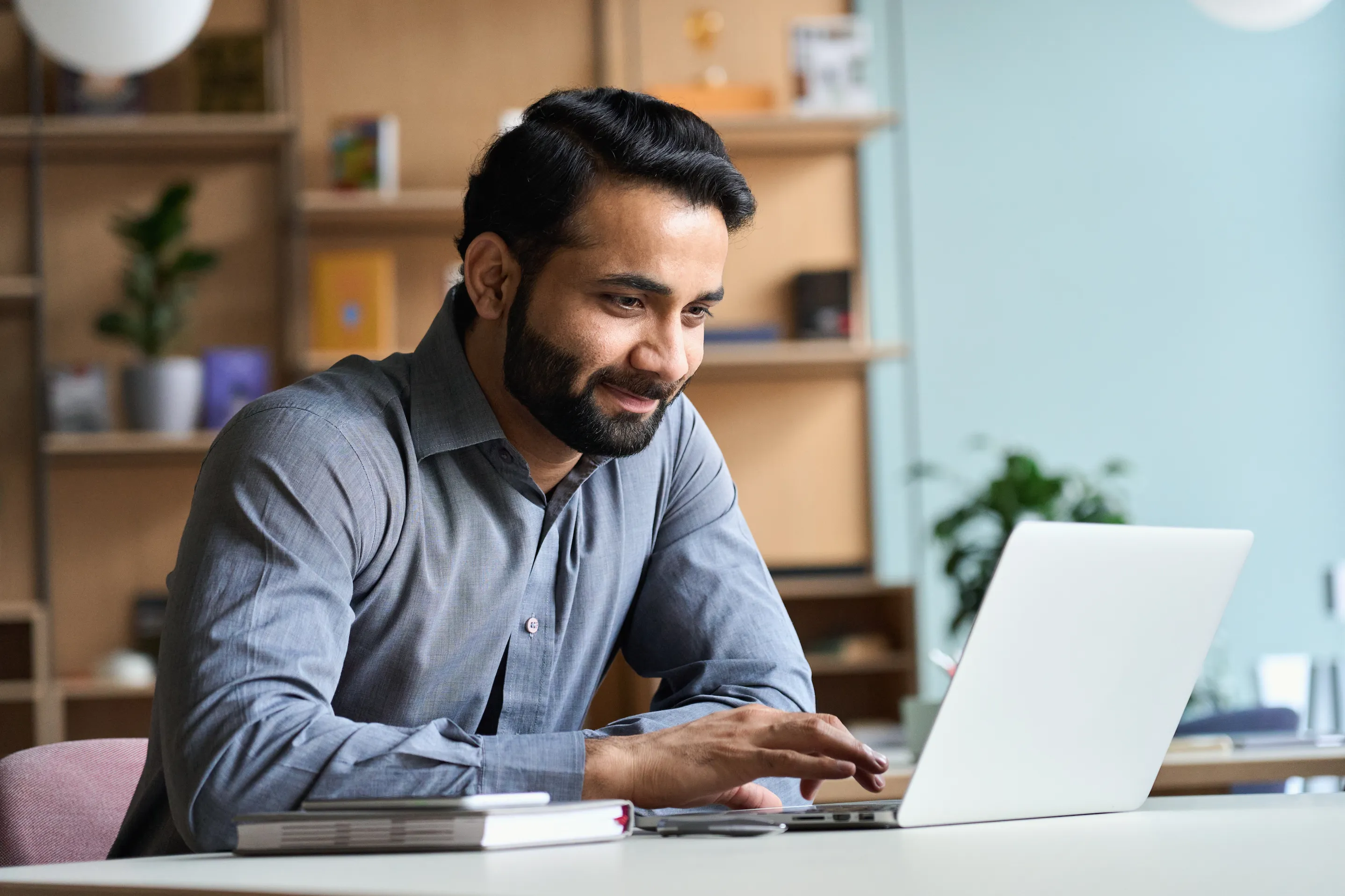 Man in a grey shirt working on a laptop at a desk, with shelves and plants in the background.