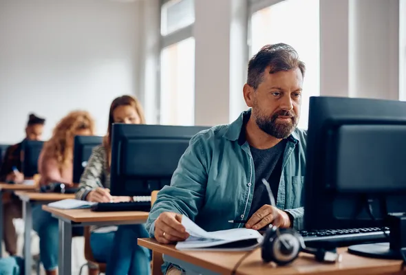 A man in a classroom setting works intently at a computer, surrounded by other students focusing on their screens and notes.