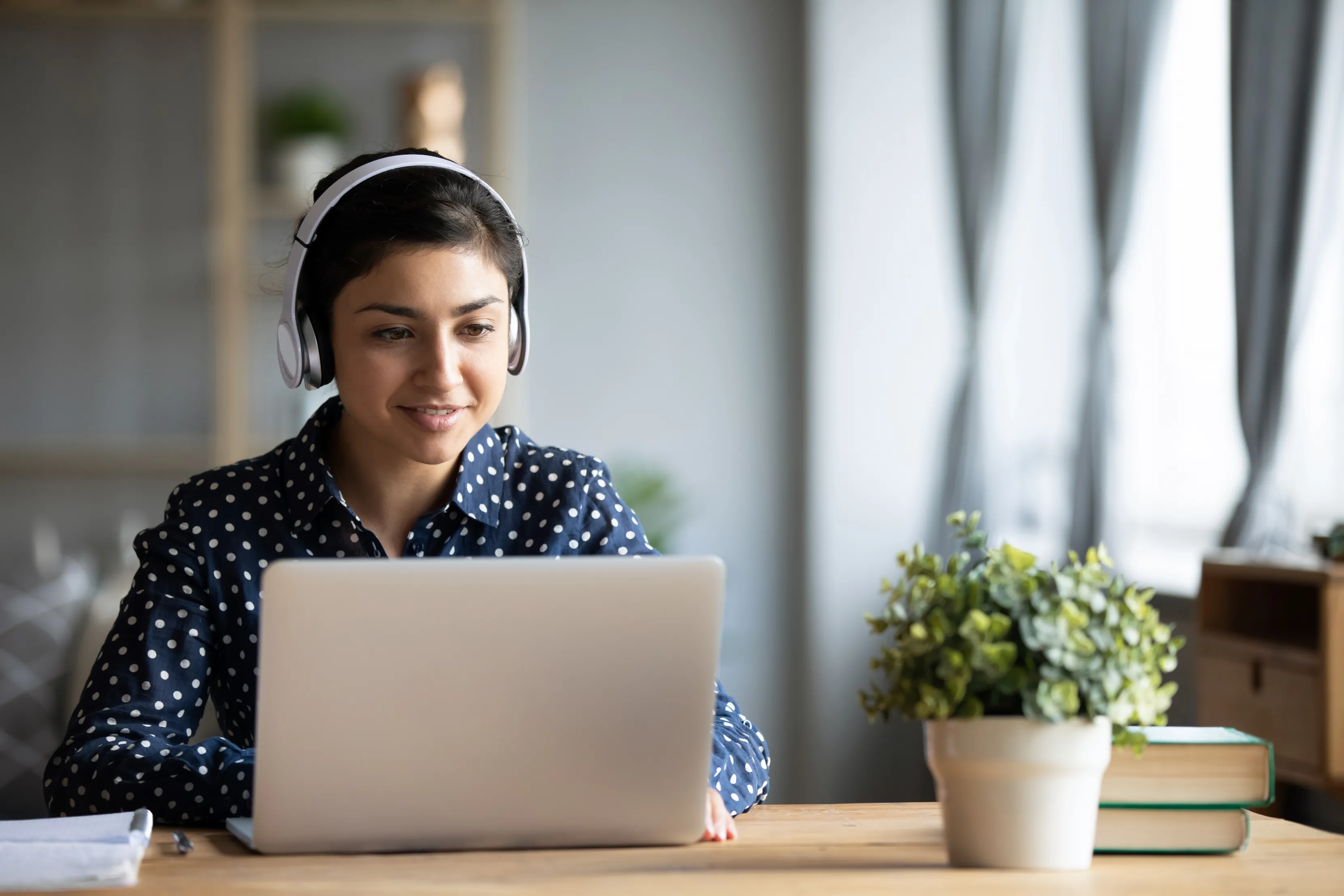 A woman wearing headphones sits at a desk, working on a laptop. A potted plant and books are nearby. She is smiling and focused.