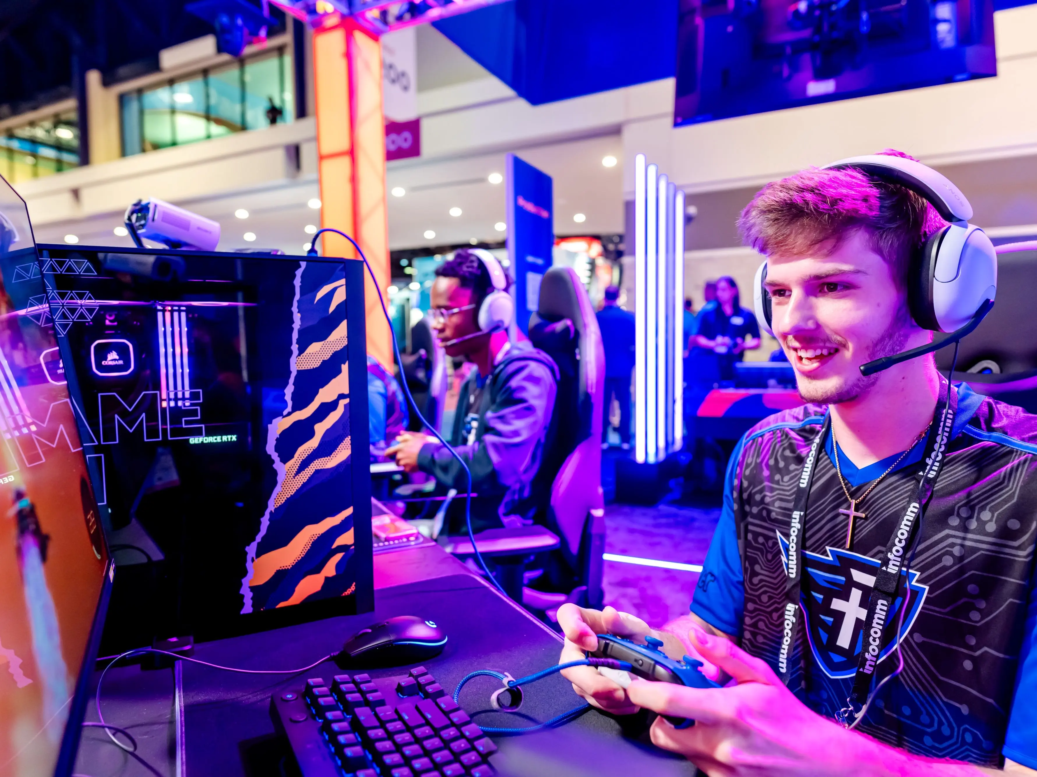 Two gamers at a gaming event playing on PCs. The gamer in the foreground is smiling, wearing headphones, and holding a controller.