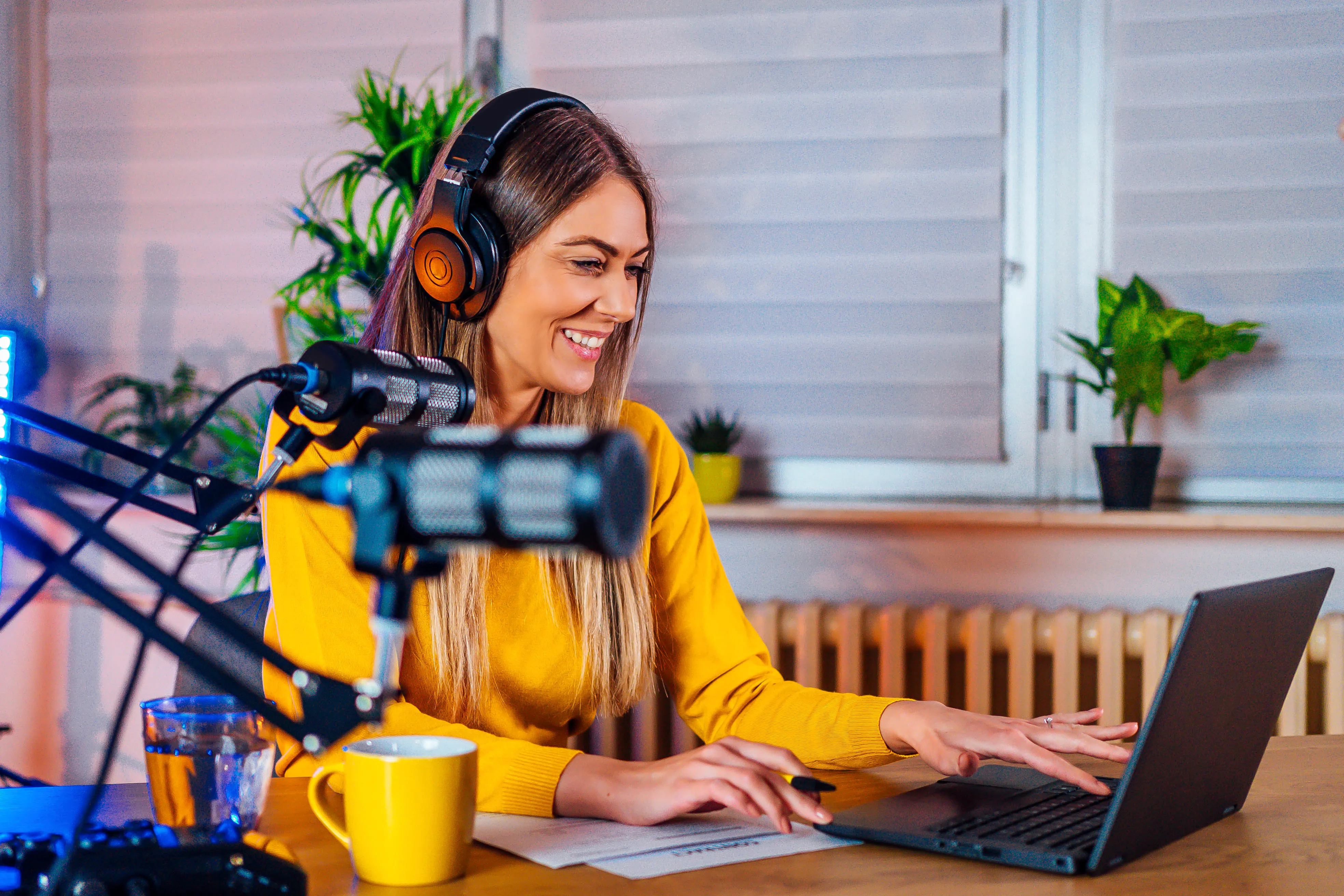 A smiling woman wearing headphones works on a laptop with a microphone nearby, in a room with potted plants and a yellow mug on the table.