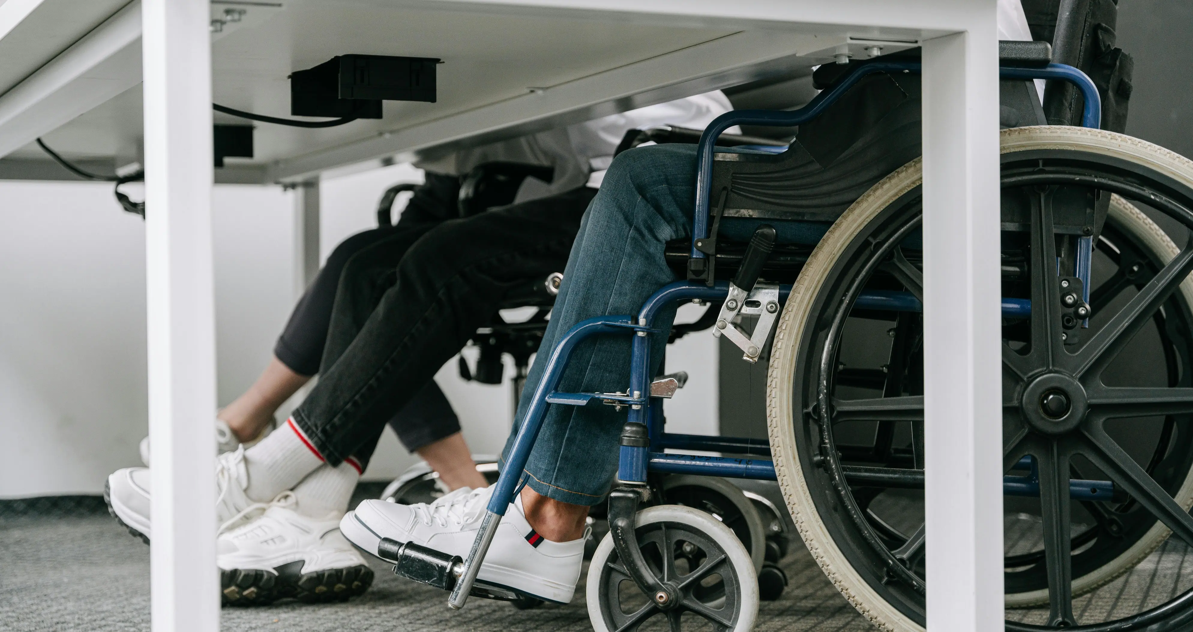 Two people in wheelchairs at a desk, legs visible in sneakers and jeans, highlighting accessibility in an office setting.