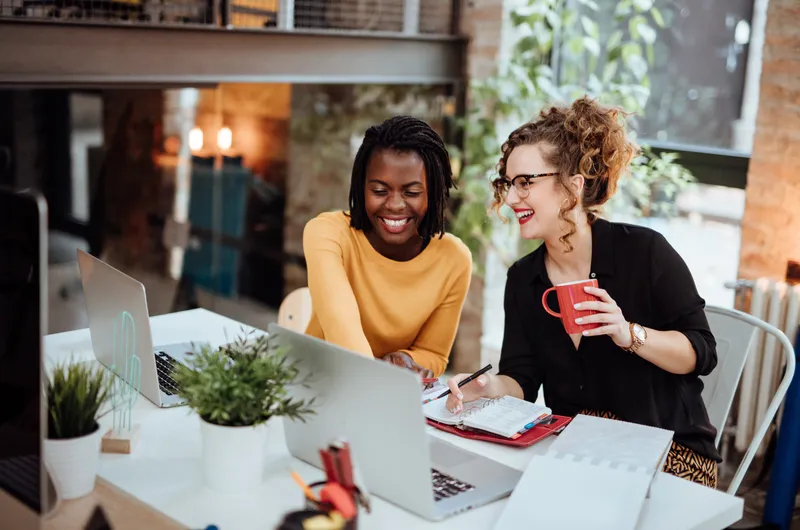 Two women smiling and collaborating at a desk with laptops, a red mug, and notebooks. Bright office space with plants.