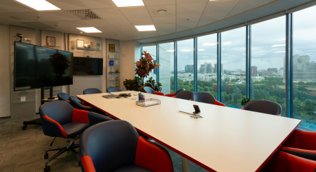 Modern conference room with a long white table, blue and red chairs, large TV screen, glass wall, and cityscape view.