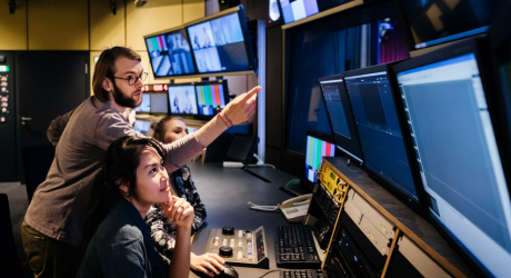 People working at a control room desk, surrounded by monitors and broadcasting equipment, focusing on video editing or production tasks.