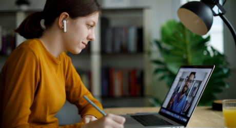 A woman in a yellow sweater wears earbuds and writes notes while watching a video call on her laptop, seated at a desk with a lamp and a glass of juice.