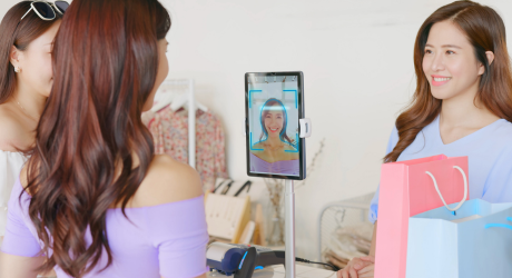 Two women at a store counter, one using facial recognition on a tablet for payment, with shopping bags visible.