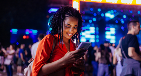 A woman in a bright orange top smiles while looking at her phone at a lively outdoor event with colorful lights in the background.