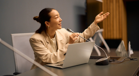 Woman speaking at a conference table, gesturing with her hand, using a laptop and microphone.