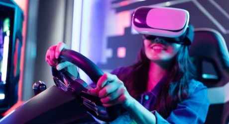 A person wearing a virtual reality headset is seated in a driving simulator, holding the steering wheel, surrounded by colorful lights.