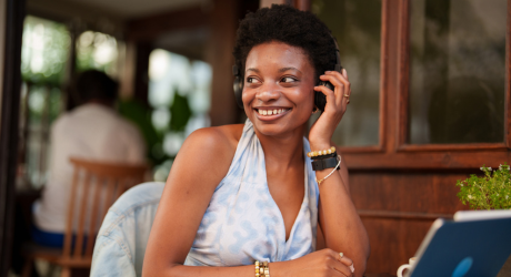 Smiling woman in a sleeveless top, on a phone call at a cafe, with a laptop and potted plant in the foreground.