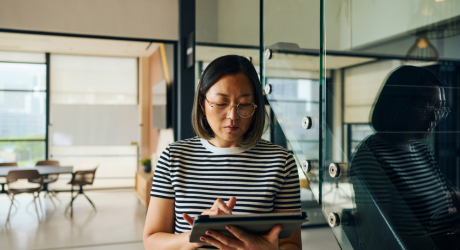 A woman with glasses and a striped shirt uses a tablet in a modern office with large windows and minimalistic decor.