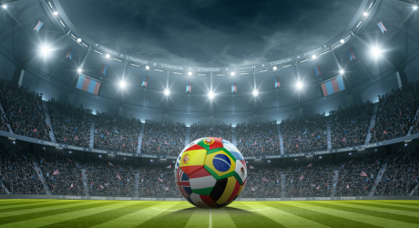 Soccer ball with various national flags on the grass in a brightly lit stadium with a cheering crowd.