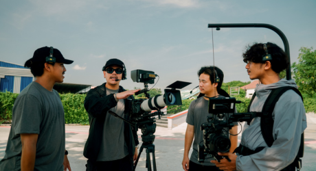 Film crew discussing a scene outdoors, with one person gesturing near a camera on a tripod, wearing headsets and casual clothing.