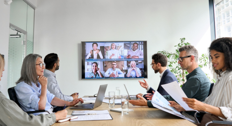 People in a conference room are having a meeting with colleagues on a screen. Papers and laptops are on the table.