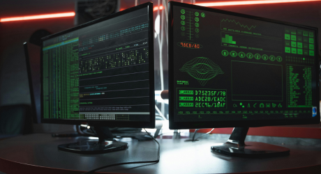 Dual-monitor setup displaying green-on-black code, system graphs, and data streams in a dark room, illustrating a hacker-style cyberwar operation or cybersecurity breach analysis.
