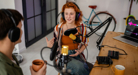 Two people recording a podcast in a casual setting, seated at a table with microphones and headphones, laptop showing audio waves.