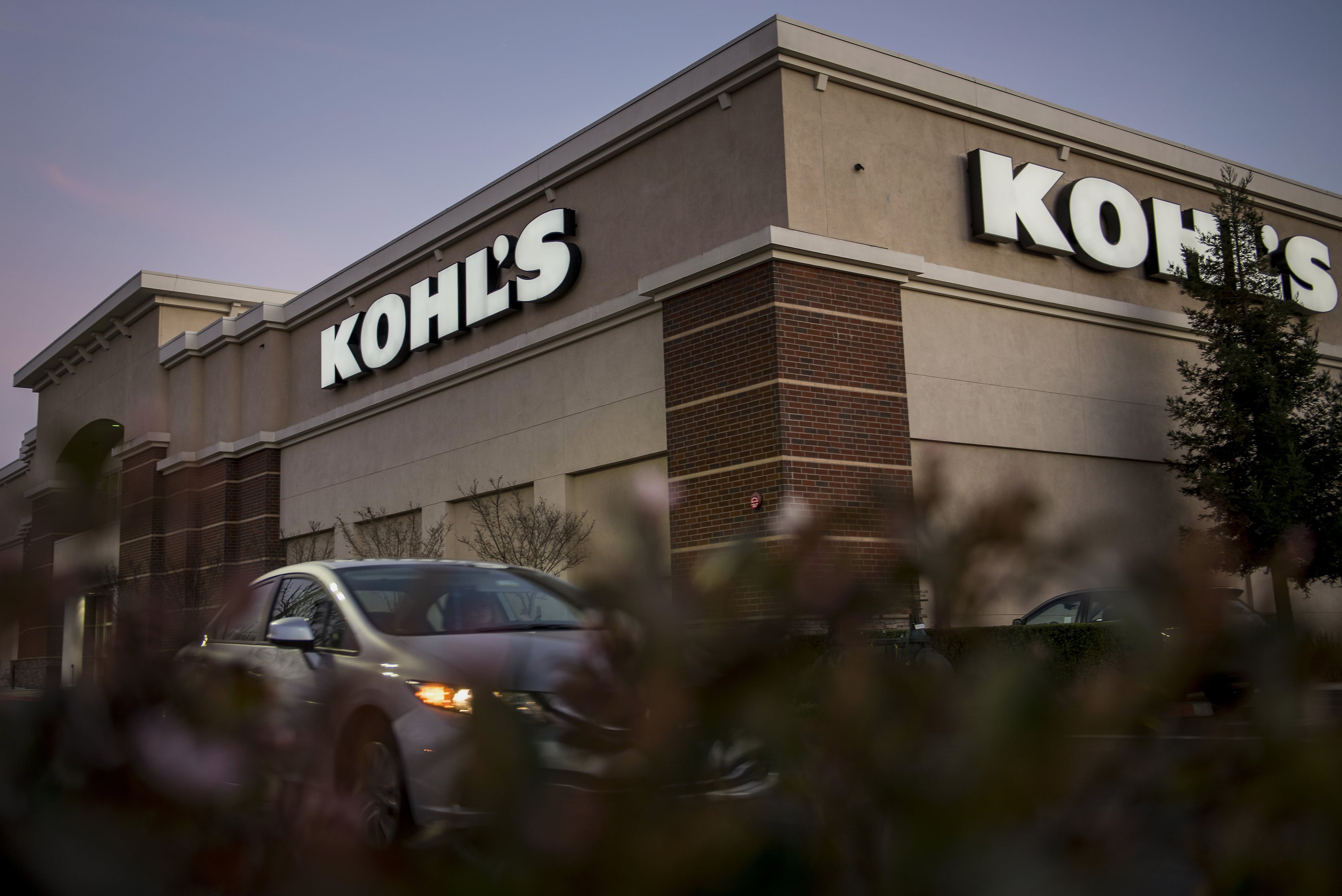 A car glides by a Kohls store during dusk, evoking a sense of the golden era. The building, with its beige and brick exterior, boasts large, illuminated Kohls signage. In the foreground, some foliage is visible, complementing this nostalgic retail scene.