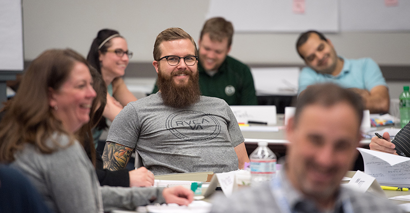 A group of people sitting at tables in a classroom setting, smiling and engaged in discussion, with a bearded man in a grey shirt in the center.