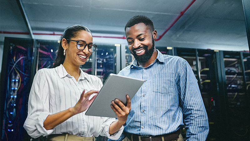 Two people smiling and discussing information on a tablet in a server room.