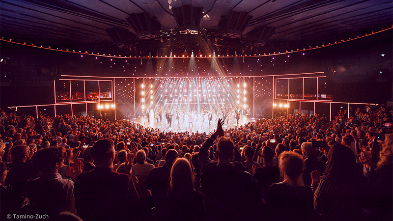 A large audience in a theater applauding performers on a brightly lit stage with spotlights and decorative vertical lights.