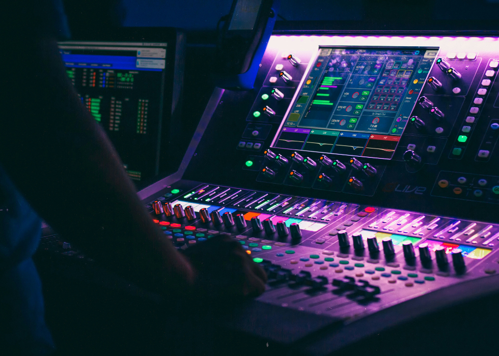 A sound and lighting control console glowing purple and blue, with faders, knobs, and a touchscreen interface being operated by a person in a dark room.