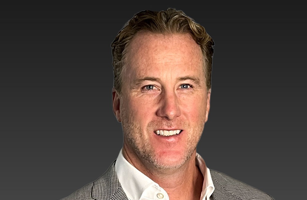 A man with short hair wearing a light gray checked blazer and white shirt, smiling against a dark gray background.