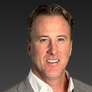 A man with short hair wearing a light gray checked blazer and white shirt, smiling against a dark gray background.