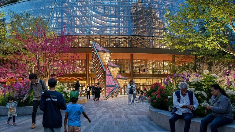 People gather outside a modern glass building surrounded by colorful trees and flowers, with a geometric sculpture at the entrance.
