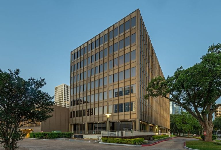 A modern multi-story office building with large windows, surrounded by trees, under a clear blue sky.