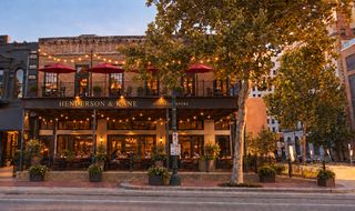 Exterior of Henderson & Kane General Store with string lights, outdoor seating, and red umbrellas, framed by large trees on a city street.