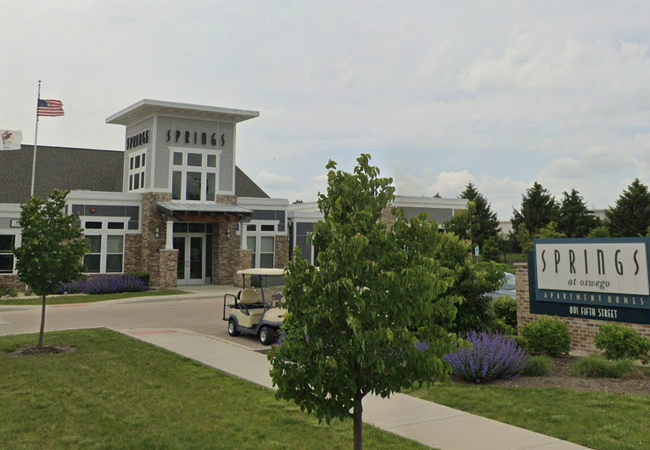 Modern building with "Springs at Oswego Apartment Homes" sign, flags, and landscaping featuring purple flowers and greenery on a sunny day.