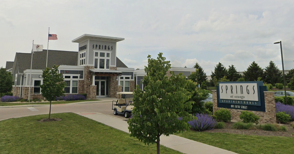 Modern building with "Springs at Oswego Apartment Homes" sign, flags, and landscaping featuring purple flowers and greenery on a sunny day.