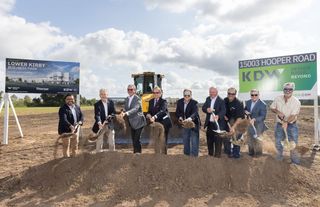 Group of men with shovels breaking ground at the Lower Kirby Business Park site, with construction equipment and signs in the background.