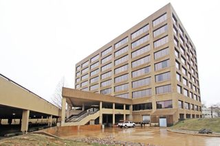A large, multi-story office building with a beige exterior and many windows, adjacent to a parking structure, on a cloudy day.