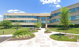 Modern three-story office building with large windows, surrounded by landscaped greenery, under a clear blue sky.