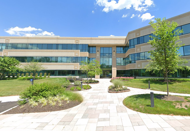 Modern three-story office building with large windows, surrounded by landscaped greenery, under a clear blue sky.