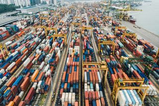 Aerial view of a busy port with numerous stacked shipping containers and large yellow cranes, surrounded by water and city buildings in the background.