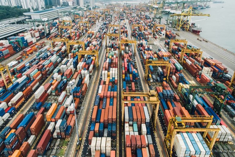 Aerial view of a busy port with numerous stacked shipping containers and large yellow cranes, surrounded by water and city buildings in the background.