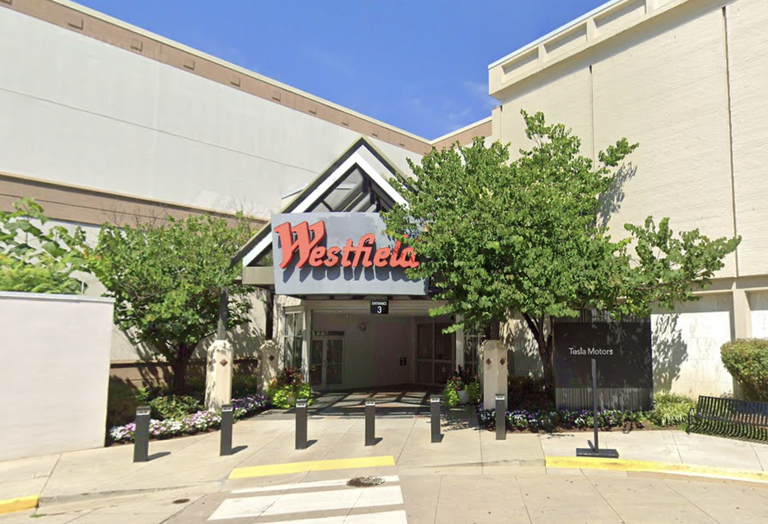 Entrance to a mall with "Westfield" signage, surrounded by trees and planters, with a Tesla Motors store visible on the right side.